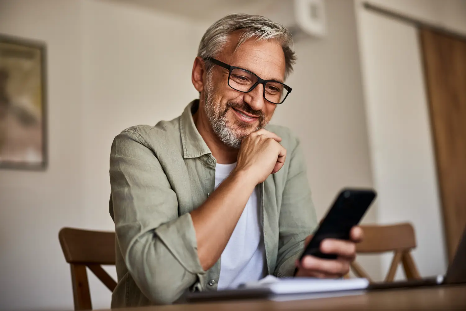 An older man with gray hair, beard, and glasses sitting at a wooden table, smiling as he looks at his phone.